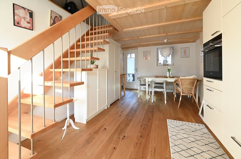 A modern, open-plan living and dining area with wooden flooring, a staircase leading to an upper level, a dining table with chairs, and a built-in oven. The space features contemporary decor, including artwork on the walls and a patterned rug.