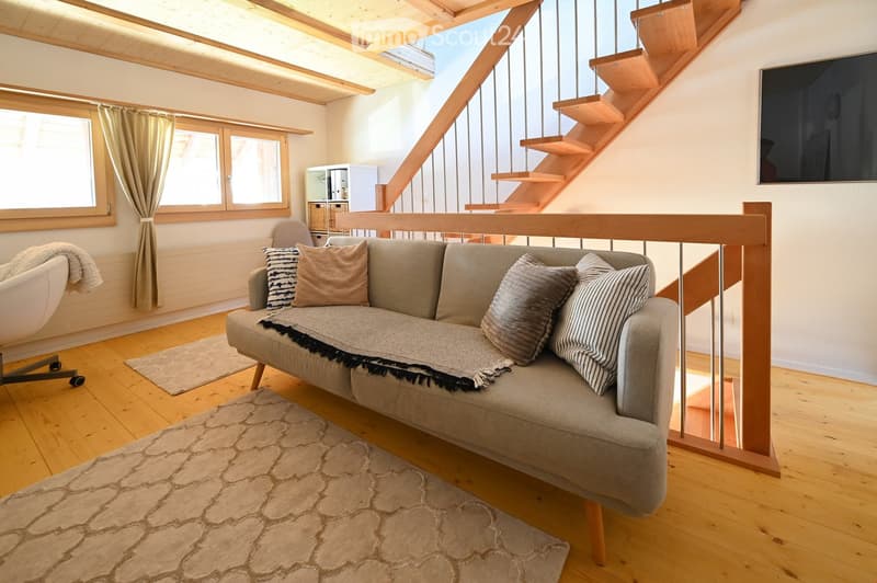 A modern living room with wooden floors, a grey sofa with decorative pillows, a white chair, a large screen, and a staircase leading upwards.