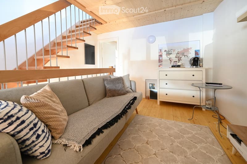 Modern living room with a grey sectional sofa, throw pillows, a coffee table, a small round table, a drawer unit with a coffee machine, and a world map on the wall. Wooden flooring and a staircase leading upwards.