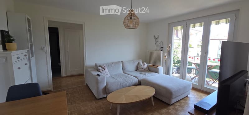 Bright living room with white walls, wooden flooring, and large sliding glass doors leading to a balcony. Features a white sectional sofa with throw pillows, a round wooden coffee table, a wooden side table, a television, and a small plant on a shelf.