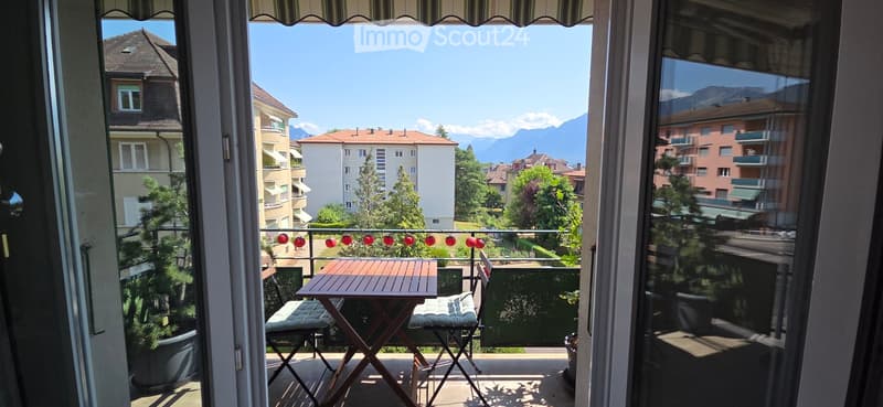 Balcony with a wooden table and two chairs, overlooking a cityscape with mountains in the background.