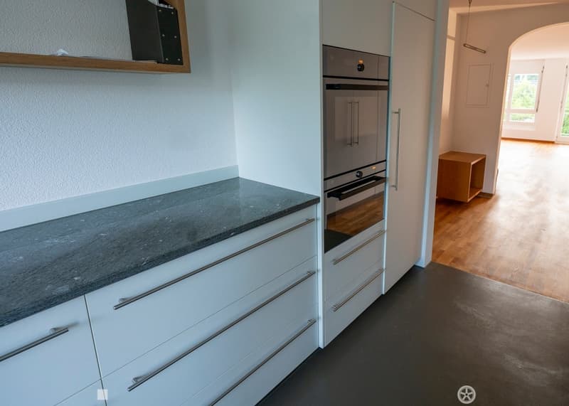 Modern kitchen with white cabinets, grey countertop, and built-in oven. Wooden floor visible in adjacent area.