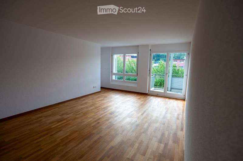 Empty room with wooden flooring, white walls, and large windows with a sliding glass door leading to a balcony.