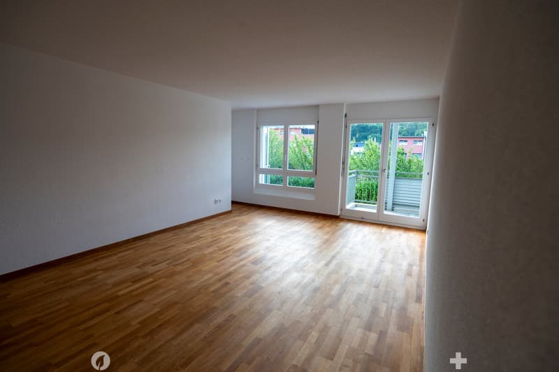 Empty room with wooden flooring, white walls, and large windows with a sliding glass door leading to a balcony.