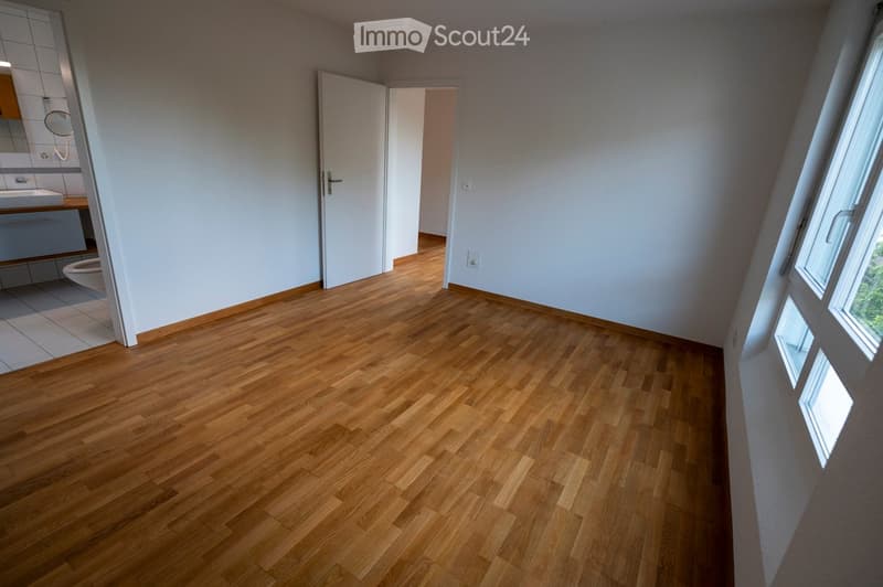Empty room with wooden flooring, white walls, and a door leading to a bathroom. The bathroom has a sink, toilet, and tiled walls.