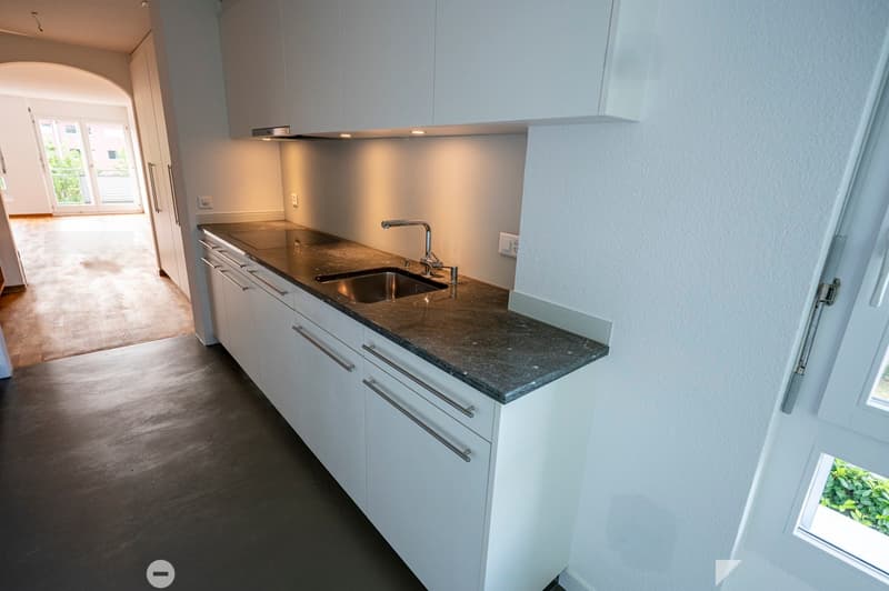 Modern kitchen with white cabinets, dark granite countertop, and a stainless steel sink. The space is well-lit with recessed lighting and has a large window providing natural light.