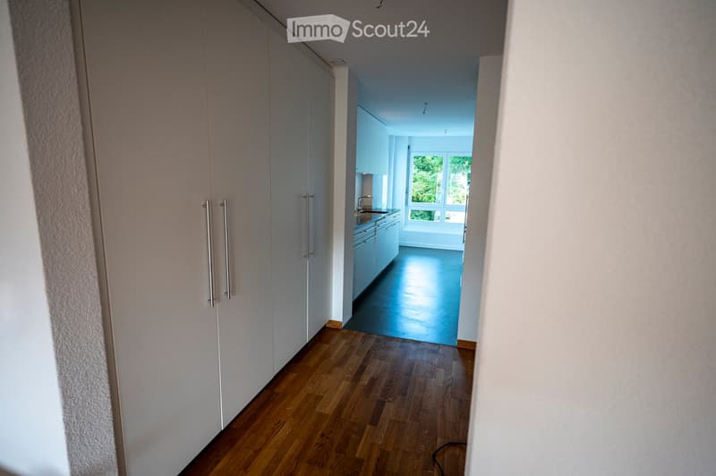 A modern hallway with wooden flooring, white walls, and built-in closet doors on one side. The hallway leads to a brightly lit kitchen area with white cabinets and a dark countertop.
