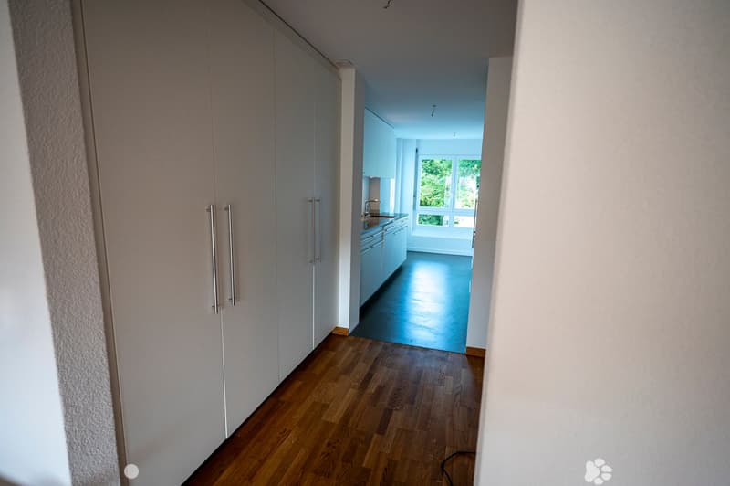 A modern hallway with wooden flooring, white walls, and built-in closet doors on one side. The hallway leads to a brightly lit kitchen area with white cabinets and a dark countertop.