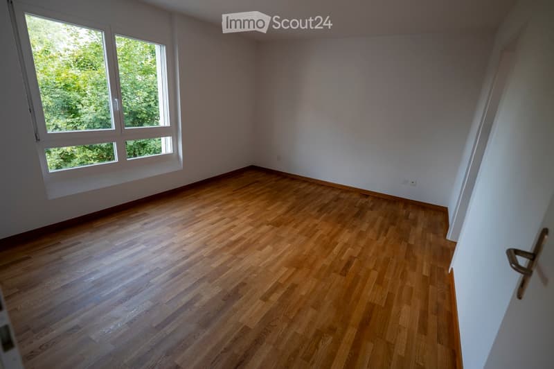 Empty room with wooden flooring and large windows showing greenery outside. No furniture or equipment present.