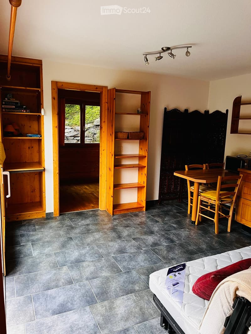 A furnished room with a sofa, a small dining table with chairs, wooden shelves, and a lamp. The room has tiled flooring and an open doorway leading to another space.
