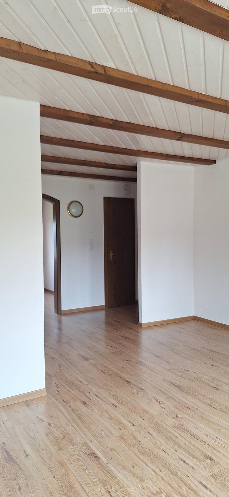 Empty room with light wooden flooring, white walls, exposed wooden beams on the ceiling, and a single door leading to another space.