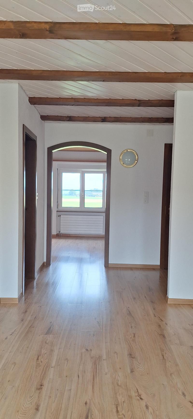 Bright, open hallway with wooden flooring, white walls, and exposed wooden beams on the ceiling. An arched doorway leads to another room with large windows and a view of the outdoors.