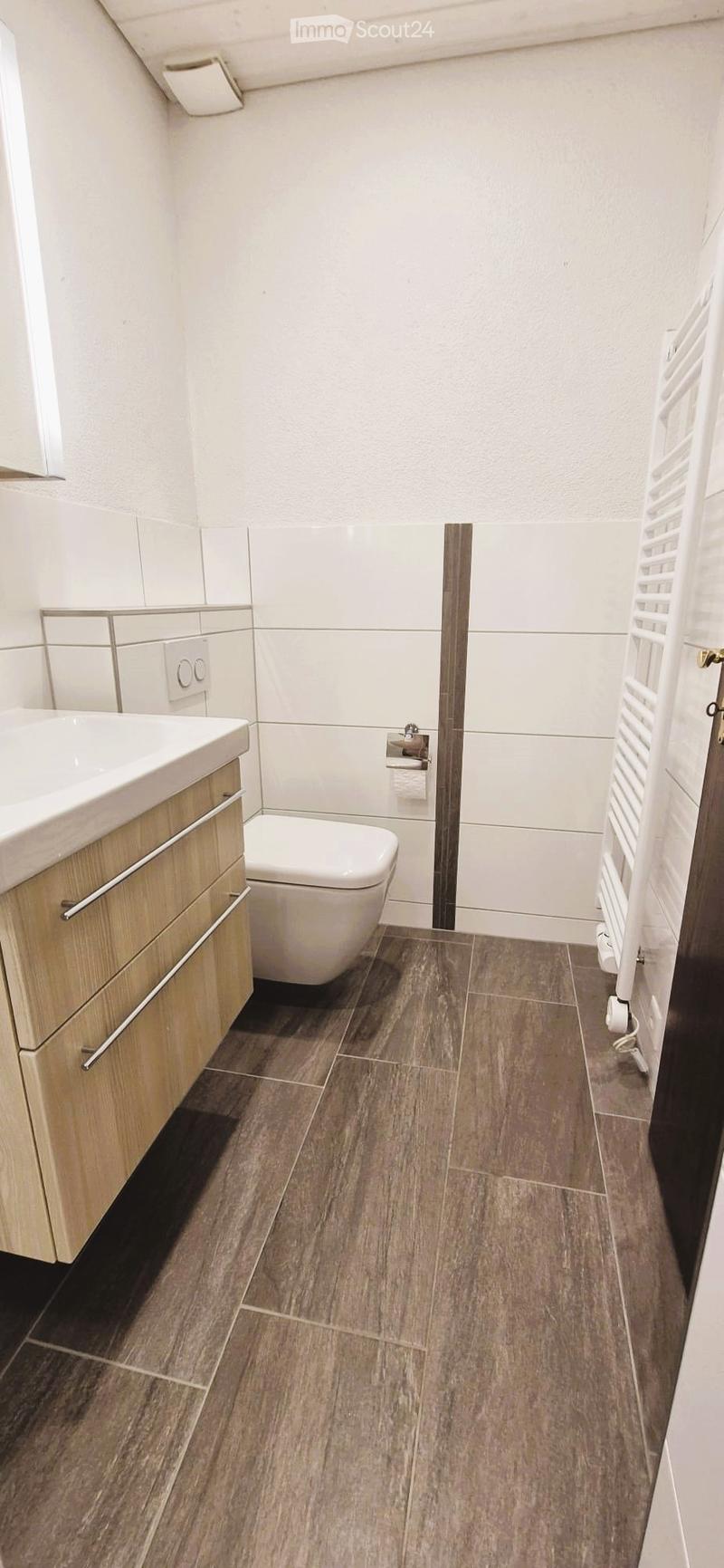 Modern bathroom with a wooden vanity, white sink, toilet, and radiator. The floor is covered with wood-effect tiles.