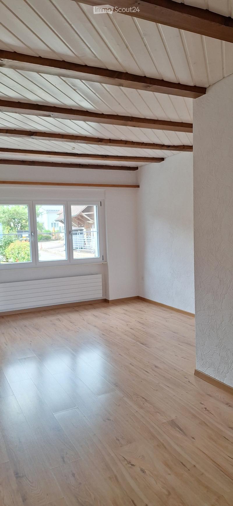 Empty room with light wooden flooring, white walls, and exposed wooden beams on the ceiling. A large window with white frames and a radiator below it.