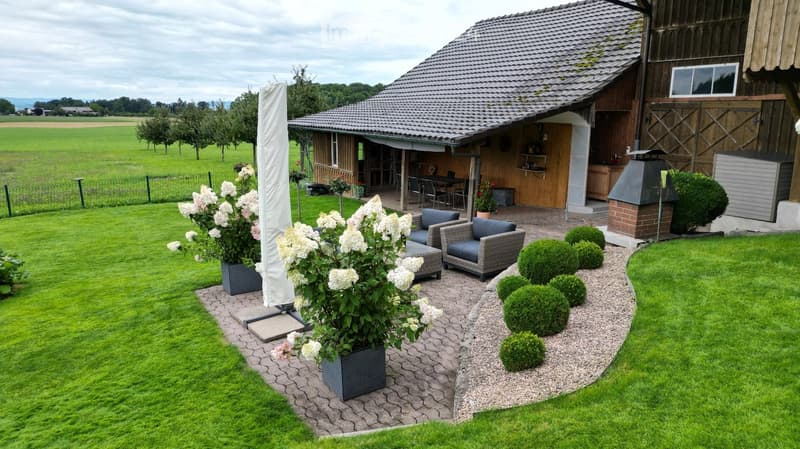 Well-maintained garden with a patio area, outdoor seating, and a barbecue area attached to a wooden structure. The lawn is neatly trimmed, and there are several potted plants and shrubs.