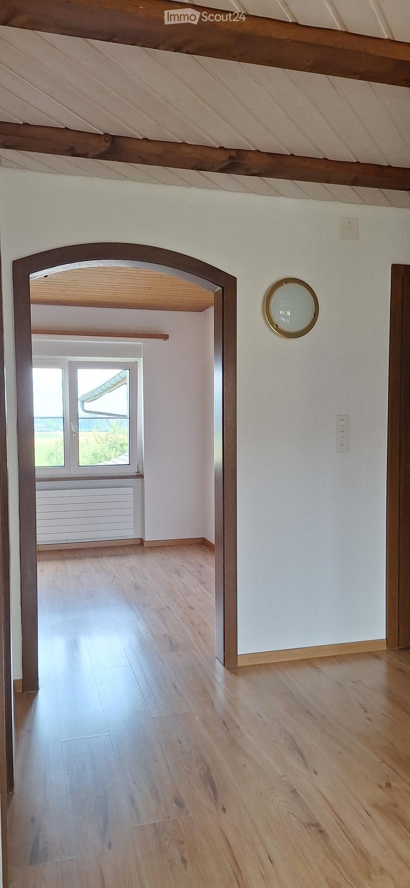 Empty room with light wooden flooring, white walls, and a large arched doorway leading to another room with a window and radiator.