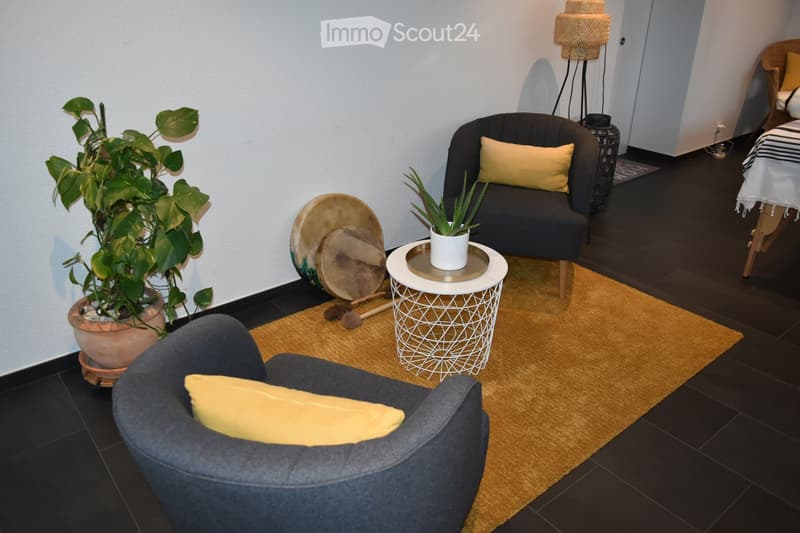 A cozy living room with dark wooden flooring, two armchairs with yellow cushions, a small white table with a plant, and a large potted plant. A drum set is placed near the wall.