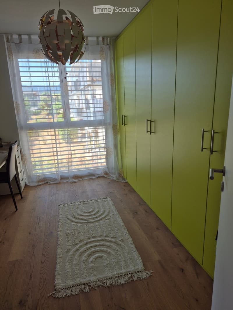 Bright, minimalist room with light green cabinets, wooden flooring, a white rug with a spiral design, and a hanging light fixture.