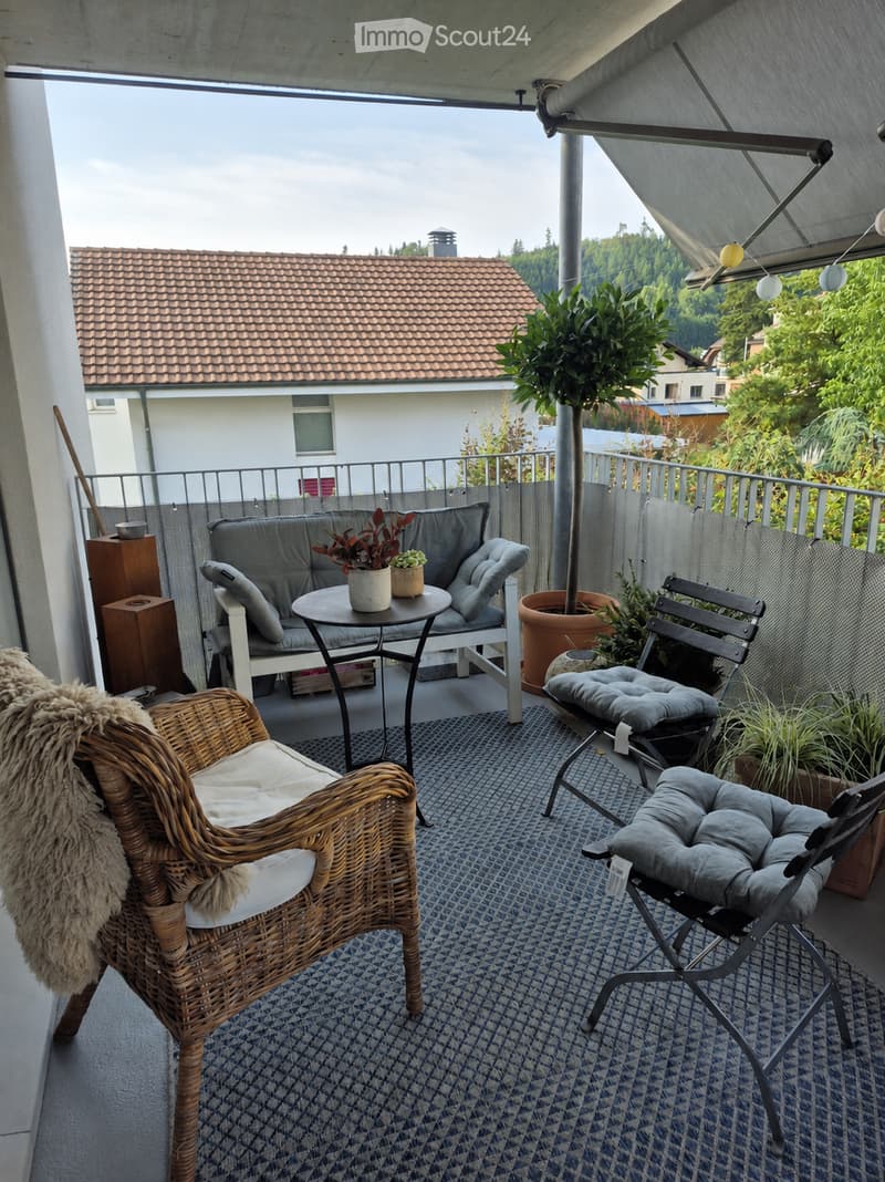 Balcony with a sofa, small round table, wicker chair, and potted plants, overlooking a residential area with trees and a house.