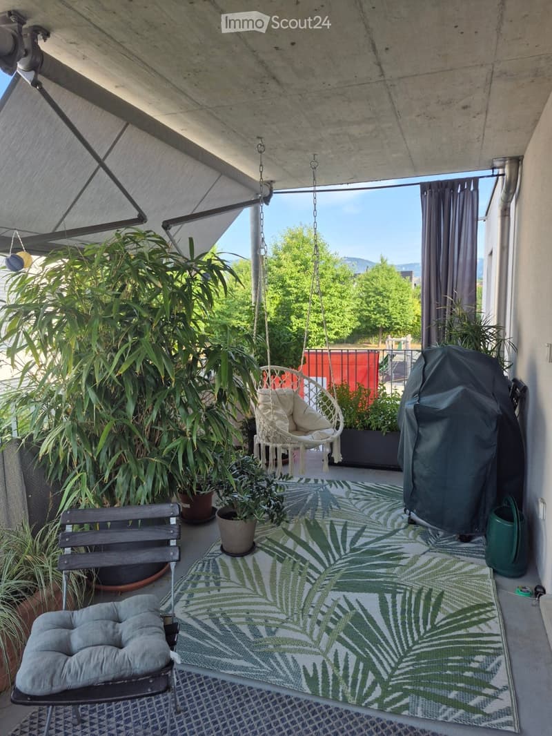 Balcony with a hanging wicker chair, a cushioned chair, a green and white palm leaf rug, and several potted plants.