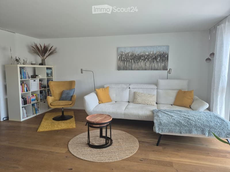 Modern living room with white walls, wooden flooring, a white sectional sofa, a yellow armchair, a small wooden table, a bookshelf, and a painting on the wall.