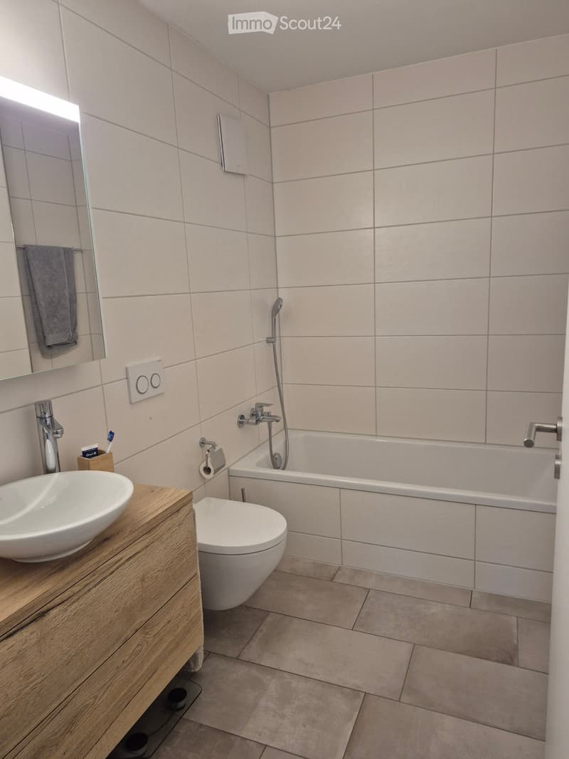Modern bathroom with a white sink, toilet, bathtub, and a mirror. The walls are tiled in light beige, and the floor is tiled in a light beige color. A wooden vanity holds the sink, and a towel hangs on a rack next to the mirror.