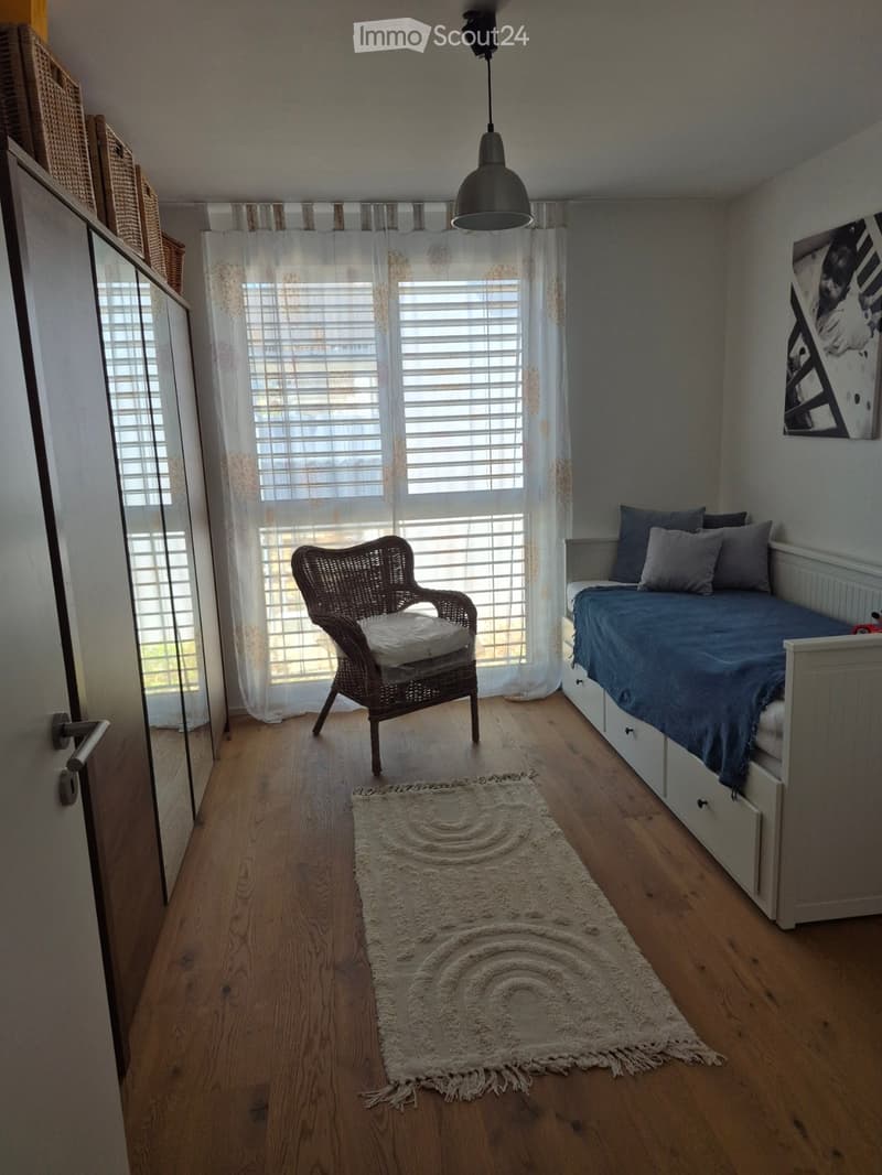 Cozy bedroom with wooden flooring, a single bed with blue bedding, a wicker chair, and a round white rug in the center.