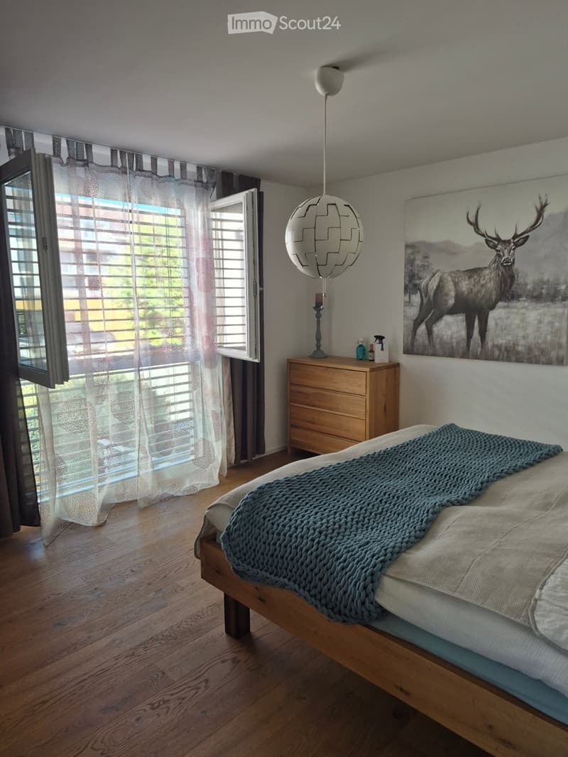 A modern bedroom with a wooden bed, a blue blanket, a nightstand with a lamp, and a large picture of a deer on the wall. The room features large windows with blinds and sheer curtains, and a hanging geometric lamp.