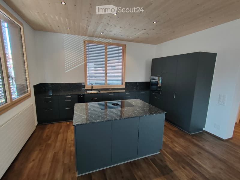 Modern kitchen with dark grey cabinets, granite countertop island, wooden flooring, and large windows with blinds.
