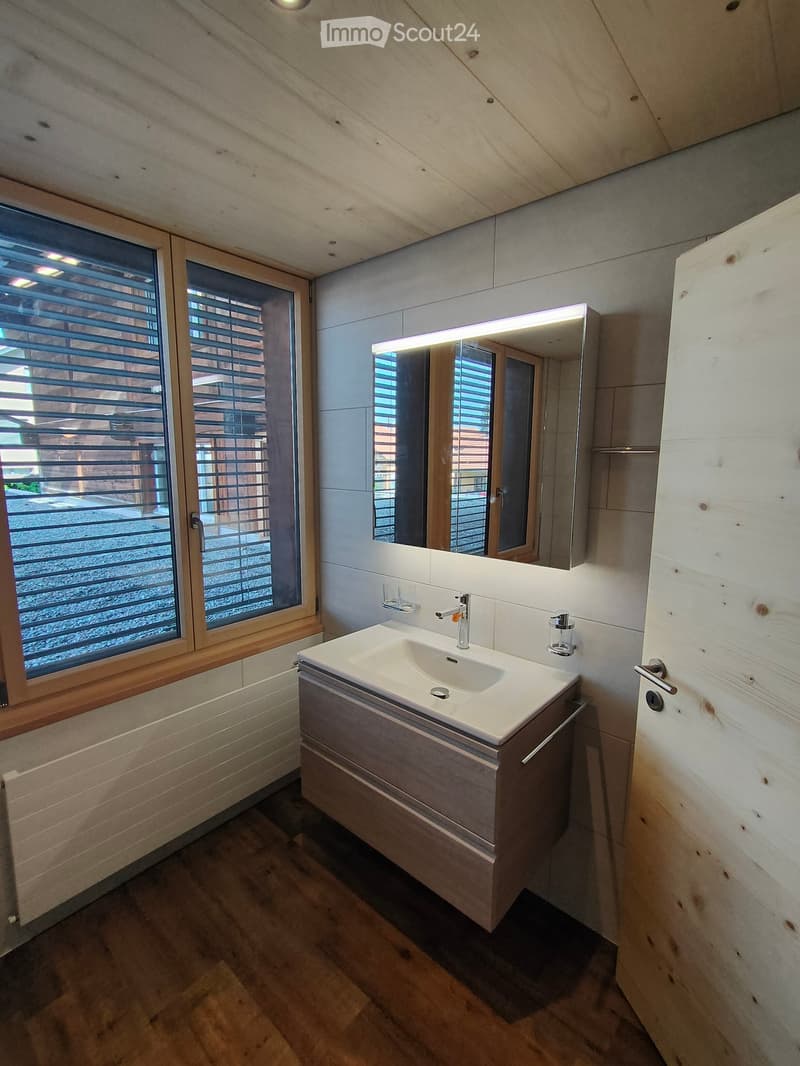 Modern bathroom with a white sink, sleek faucet, illuminated mirror, and wooden cabinet. Large window with blinds provides natural light.