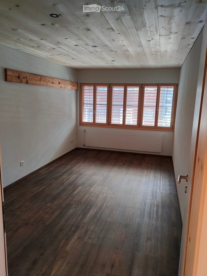 Empty room with wooden flooring, white walls, large windows with blinds, and a radiator under the windows.