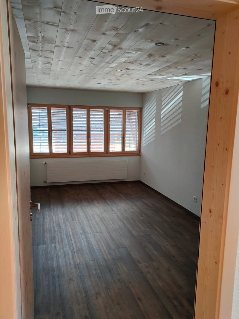 Empty room with wooden flooring, white walls, large windows with blinds, and a radiator under the windows.
