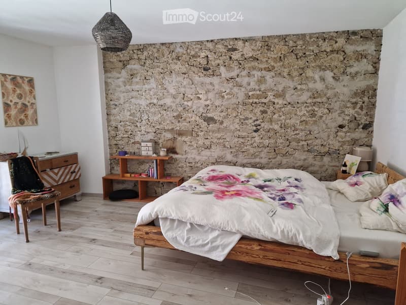 Cozy bedroom with a stone accent wall, wooden bed frame, floral bedding, and a mix of modern and rustic furniture including a wooden nightstand, a wooden shelf, a wooden dresser, and a hanging woven lamp.