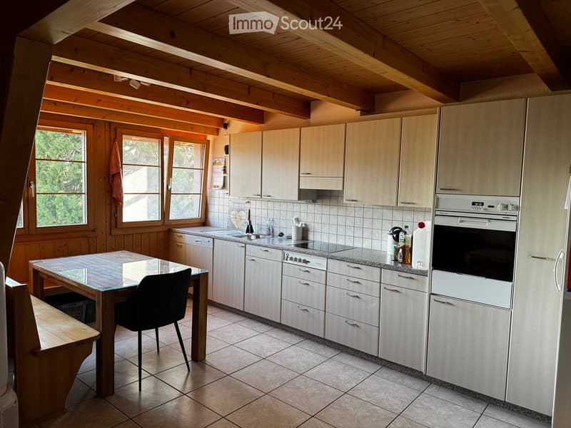 Bright kitchen with wooden beams, white cabinets, granite countertops, and a central island table with a black chair. Appliances include an oven, dishwasher, and refrigerator. Natural light streams in through large windows.