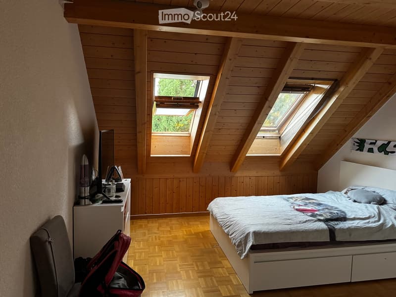 Cozy attic bedroom with wooden walls and ceiling, skylight windows, a bed with a duvet, a nightstand with a laptop, and a chair with a backpack nearby.