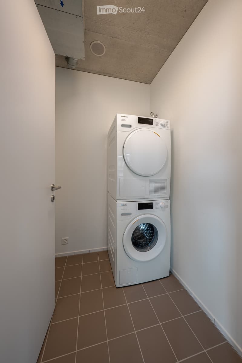 A small bathroom with a stacked washing machine and dryer unit, tiled floor, white walls, and a door.
