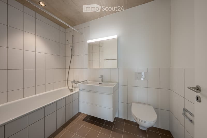 Modern bathroom with white tiles, a bathtub, a sink with a modern faucet, a toilet, and a mirrored cabinet with built-in lighting.