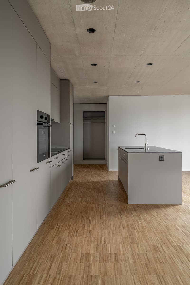 Modern kitchen with light grey cabinets, wooden flooring, and a central island with a sink and faucet. An oven is visible on the left side.