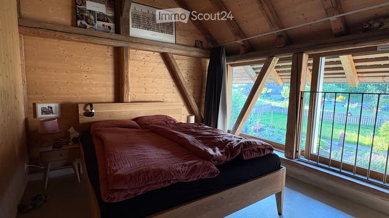 Cozy attic bedroom with wooden walls, a large window offering a view of the garden, a neatly made bed with a pink duvet, a nightstand with a lamp, and framed pictures on the wall.