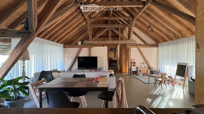 Open-concept living and dining area with exposed wooden beams, large windows, a sofa, dining table, chairs, television, and decorative items.