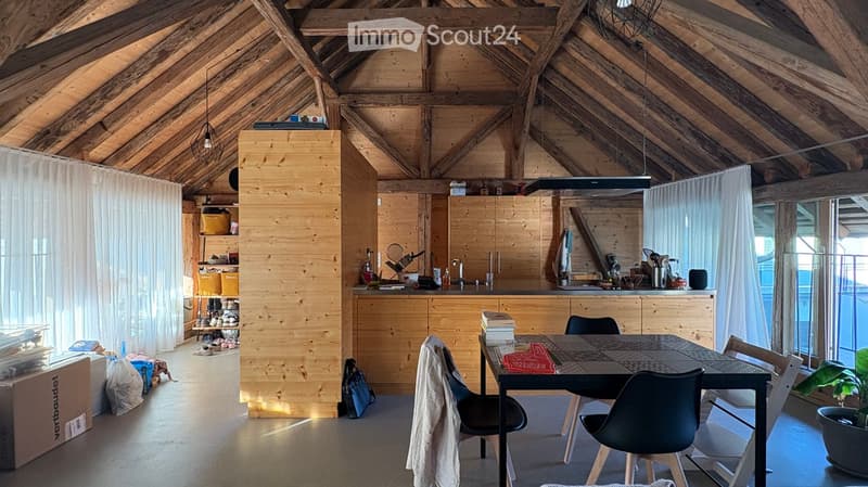 Open-concept room with exposed wooden beams, a dining table with chairs, a kitchen area with cabinets and a counter, and a curtained area with shelves.