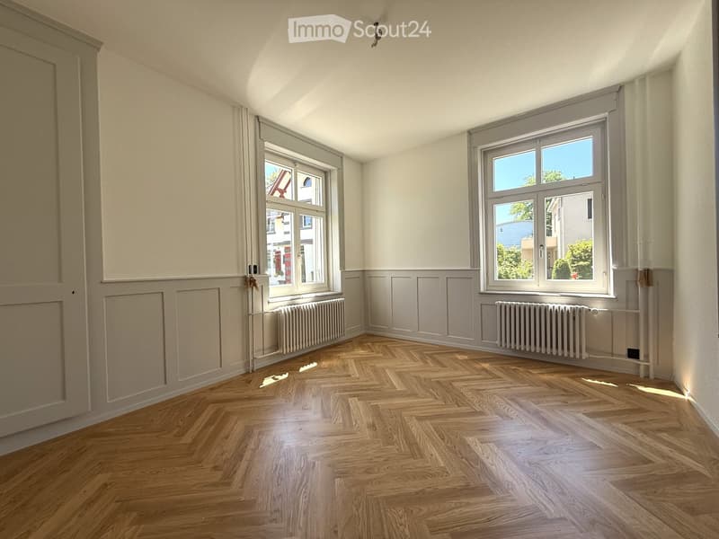 Empty room with wooden herringbone flooring, white walls, and two windows with radiators beneath them.