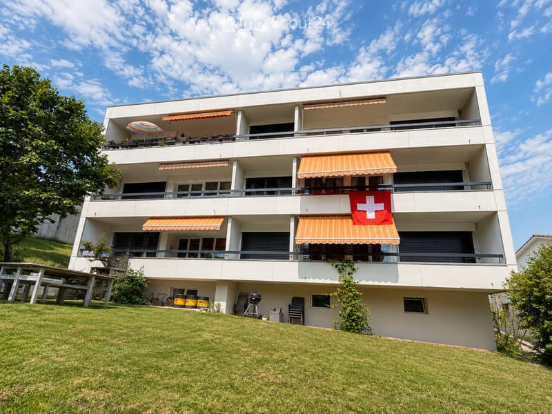 Modern three-story apartment building with balconies, orange awnings, and a Swiss flag. The building is surrounded by a well-maintained lawn and features outdoor furniture and a barbecue area.