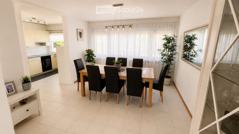 Bright dining area with a wooden dining table and six black chairs, surrounded by potted plants and a modern kitchen area with white cabinets and an oven.