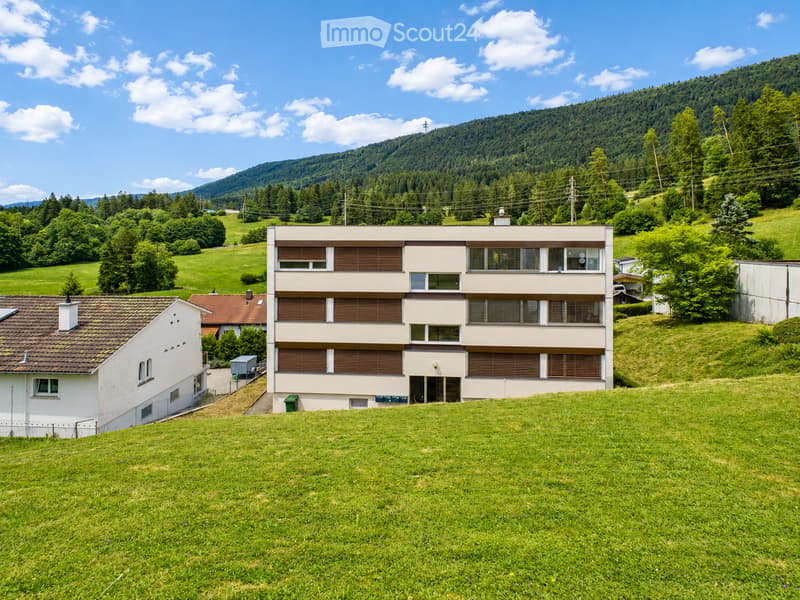 A modern two-story building with beige walls and brown shutters, situated on a grassy hillside with surrounding trees and mountains in the background.