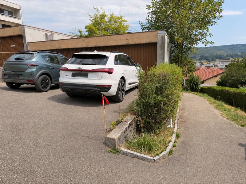 Two cars parked in a driveway next to a modern building with a concrete facade and wooden paneling. The driveway is bordered by greenery and a small pathway leads to the building.
