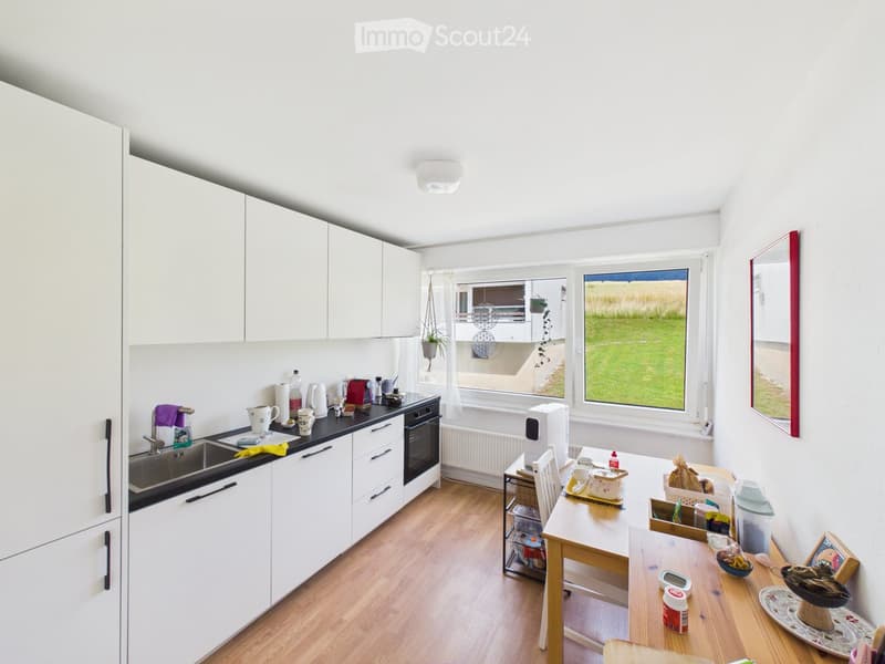 Bright, modern kitchen with white cabinets, wooden flooring, and a large window overlooking a green field. The kitchen features a sink, oven, refrigerator, and a small dining area with a table and chairs.