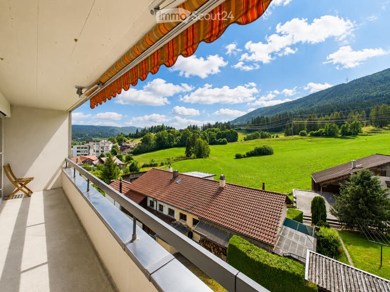 Balcony with a view of a green valley, mountains, and houses. Features a wooden chair and an awning.