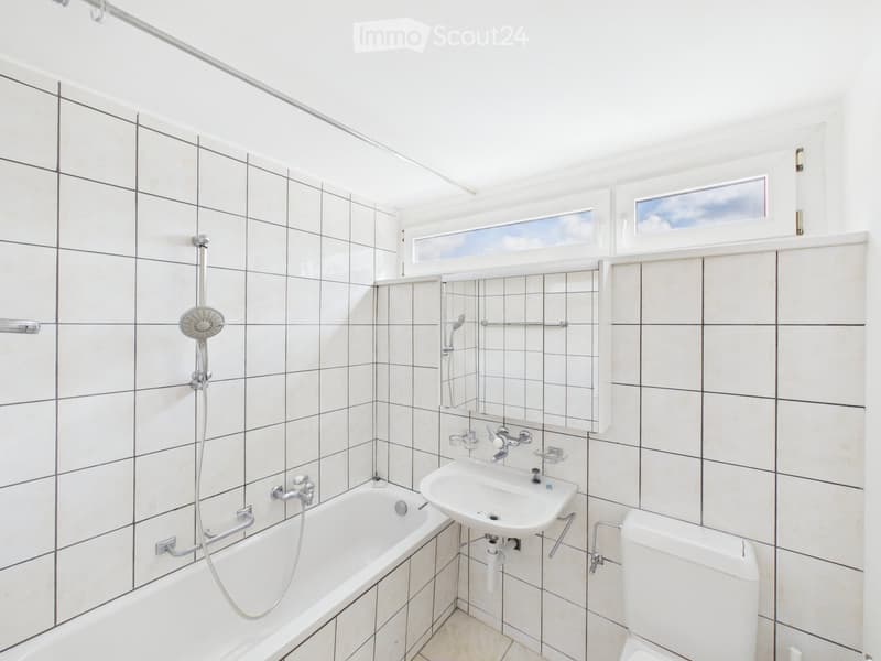 Modern bathroom with white tiled walls and floor, featuring a bathtub, sink, toilet, mirror, and shower head.