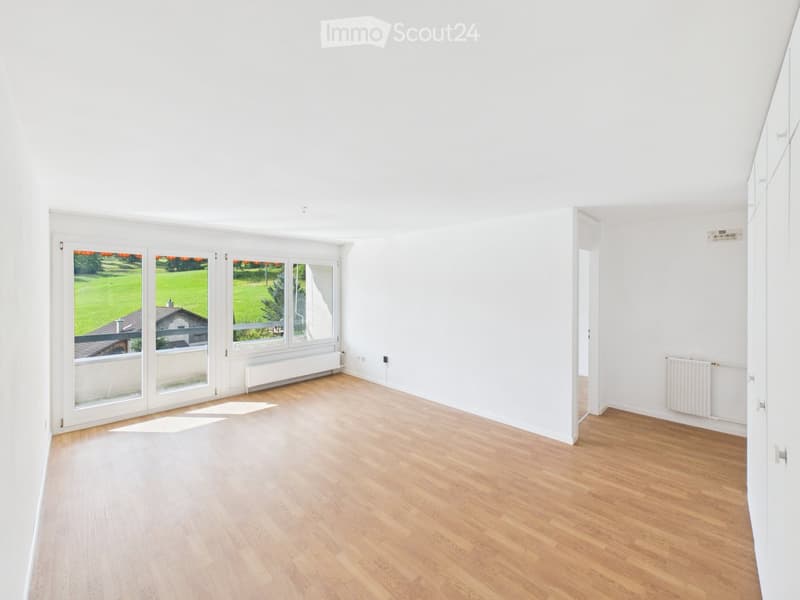 Empty room with light wooden flooring, white walls, large windows with a view of a green area, and radiators on the walls.